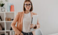 Portrait of successful business woman with glasses and in light jacket smiling against white office.