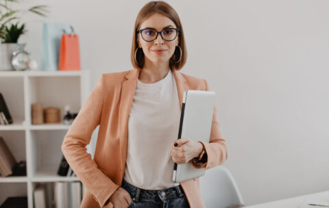 Portrait of successful business woman with glasses and in light jacket smiling against white office.