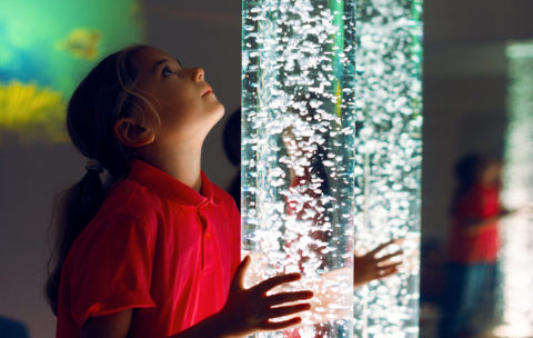 Child in therapy sensory stimulating room, snoezelen. Child interacting with colored lights bubble tube lamp during therapy session.