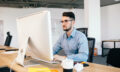 Young dark-haired man is working with  computer  at his desktop in office. He wears blue shirt and looks busy