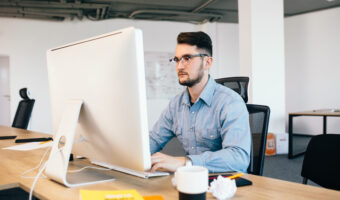 Young dark-haired man is working with  computer  at his desktop in office. He wears blue shirt and looks busy