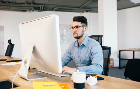 Young dark-haired man is working with  computer  at his desktop in office. He wears blue shirt and looks busy