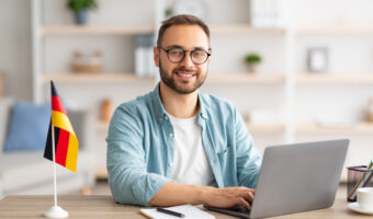 Happy young guy sitting at table with flag of Germany, using laptop pc, studying foreign language online at home