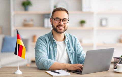 Happy young guy sitting at table with flag of Germany, using laptop pc, studying foreign language online at home