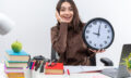 excited-young-school-woman-sits-table-with-school-tools-holding-wall-clock-putting-hand-cheek