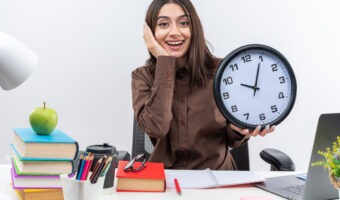 excited-young-school-woman-sits-table-with-school-tools-holding-wall-clock-putting-hand-cheek
