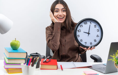 excited-young-school-woman-sits-table-with-school-tools-holding-wall-clock-putting-hand-cheek