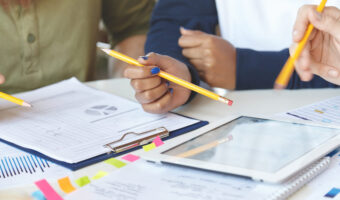 Cropped view of students working on collaborative project at canteen, making research, studying statistics and diagrams on electronic tablet, making changes and amendmends in notes using pencils
