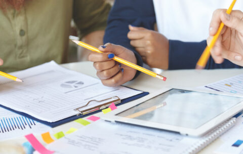 Cropped view of students working on collaborative project at canteen, making research, studying statistics and diagrams on electronic tablet, making changes and amendmends in notes using pencils