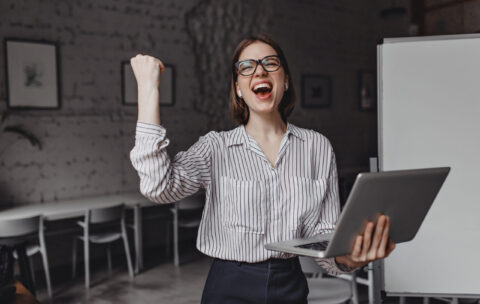Girl happily screams and makes winning hand gesture, holding laptop and posing in office against background of board.