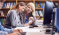 Female University Or College Student Working At Computer In Library Being Helped By Tutor