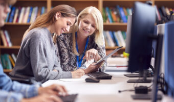 Female University Or College Student Working At Computer In Library Being Helped By Tutor