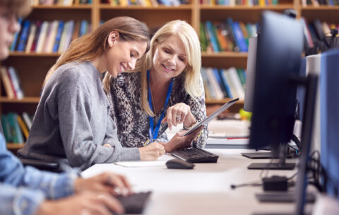 Female University Or College Student Working At Computer In Library Being Helped By Tutor