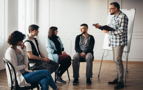Handsome senior professional therapist stands in front of a group of people of different ages during meeting for people with social problems