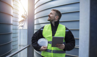 Portrait of factory worker with tablet computer standing on metal platform between industrial storage tanks and looking sideways.