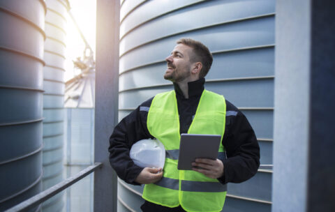 Portrait of factory worker with tablet computer standing on metal platform between industrial storage tanks and looking sideways.