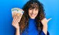 Young hispanic girl holding bunch of 50 euro banknotes pointing thumb up to the side smiling happy with open mouth