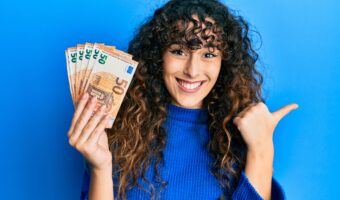 Young hispanic girl holding bunch of 50 euro banknotes pointing thumb up to the side smiling happy with open mouth