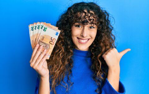 Young hispanic girl holding bunch of 50 euro banknotes pointing thumb up to the side smiling happy with open mouth